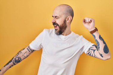 Young hispanic man with tattoos standing over yellow background dancing happy and cheerful, smiling moving casual and confident listening to music 