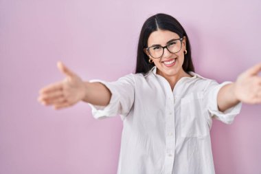 Young brunette woman standing over pink background looking at the camera smiling with open arms for hug. cheerful expression embracing happiness. 