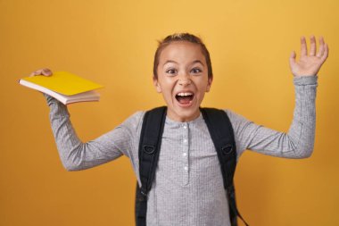 Küçük beyaz çocuk öğrenci çantası takıyor ve elinde zaferi kutlayan bir kitap tutuyor mutlu bir gülümsemeyle ve ellerini kaldırarak kazanan bir ifadeyle. 