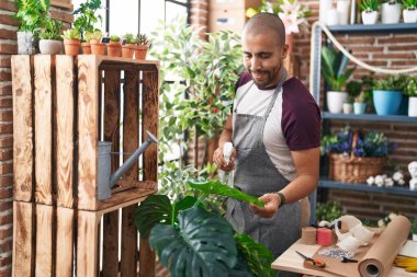 Young latin man florist using diffuser watering plant at flower shop
