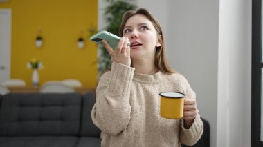 Young blonde woman talking on smartphone drinking coffee at home