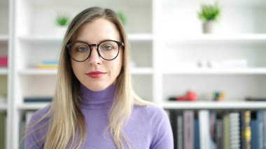 Young blonde woman student standing with relaxed expression at library university