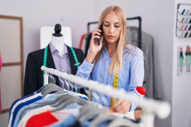 Elbiseleri askıda tutan genç sarışın kadın terzide akıllı telefondan konuşuyor.