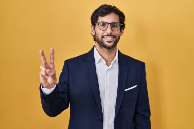 Sarı arka planda dikilen yakışıklı Latin adam iki numaralı parmaklarıyla işaret ederken kendinden emin ve mutlu gülümsüyor.. 