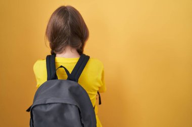 Sarı arka planda öğrenci çantası takan beyaz bir kadın. Kollarını kavuşturup arkasını kollarını çeviriyor. 