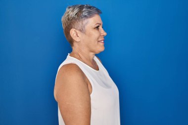 Orta yaşlı, beyaz bir kadın mavi arka planda duruyor. Kendine güveni tam, rahat bir görünüm takınıyor.. 