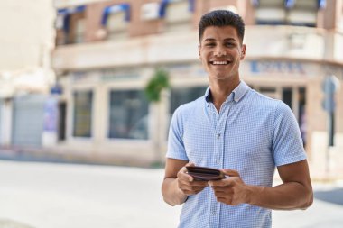 Young hispanic man smiling confident holding leather wallet at street