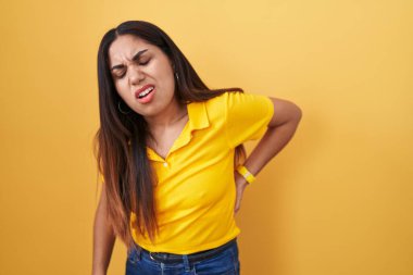 Sarı arka planda duran genç Arap kadın sırt ağrısı çekiyor, elleriyle dokunuyor, kas ağrısı çekiyor. 