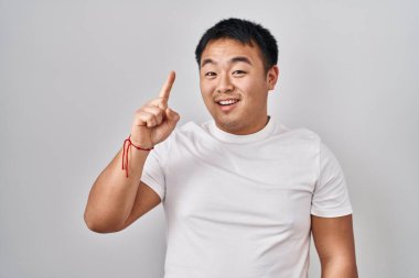 Beyaz arka planda duran genç Çinli adam başarılı bir fikirle işaret ediyor. Heyecanlı ve mutlu. Bir numara.. 