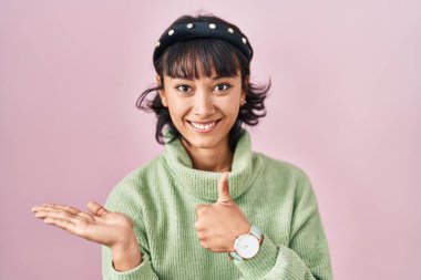 Young beautiful woman standing over pink background showing palm hand and doing ok gesture with thumbs up, smiling happy and cheerful 