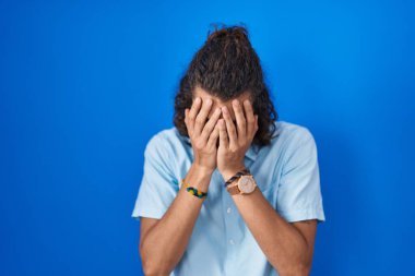 Mavi arka planda duran genç İspanyol adam. Ağlarken suratını ellerle kapatan üzgün bir ifade. depresyon kavramı. 