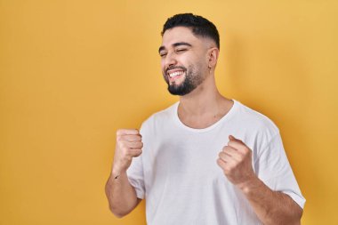 Sarı arka plan üzerine gündelik tişört giyen yakışıklı genç adam mutlu ve heyecanlı bir şekilde kollarını kaldırıp, gülümseyerek ve başarı için bağırarak kazanan jestler yapıyor. kutlama konsepti. 