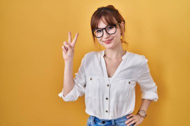 Sarı arka planda rahat bir tişört giyen genç ve güzel bir kadın kameraya bakıp zafer işareti yapıyor. İki numara.. 