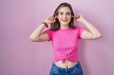 Blonde caucasian woman standing over pink background smiling pulling ears with fingers, funny gesture. audition problem 