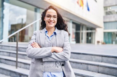 Genç İspanyol kadın yönetici gülümsüyor. Kollarını kavuşturup caddede duruyor.