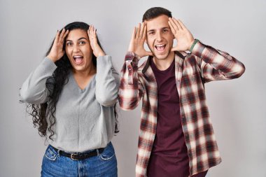 Young hispanic couple standing over white background smiling cheerful playing peek a boo with hands showing face. surprised and exited 