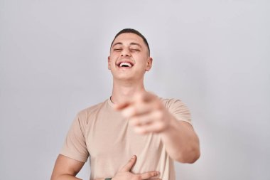 Young man standing over isolated background laughing at you, pointing finger to the camera with hand over body, shame expression 