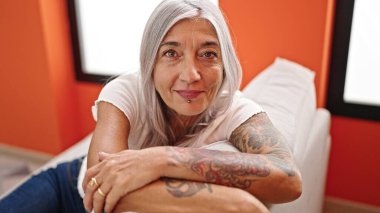 Orta yaşlı, kır saçlı, gülümseyen bir kadın. Evdeki kanepede oturuyor.