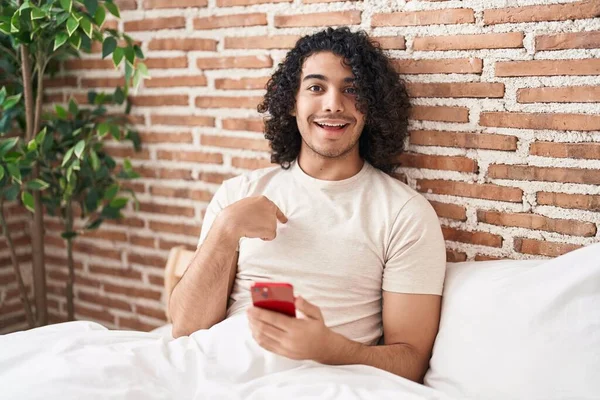 Ισπανόφωνος Άνθρωπος Σγουρά Μαλλιά Χρησιμοποιώντας Smartphone Κάθεται Στο Κρεβάτι Δείχνοντας — Φωτογραφία Αρχείου