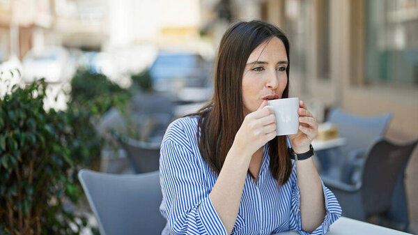 Young beautiful hispanic woman drinking coffee sitting on table at coffee shop terrace