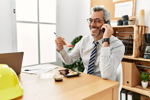 中年白髪の建築家がオフィスで寿司を食べるスマートフォンについて語る — ストック写真