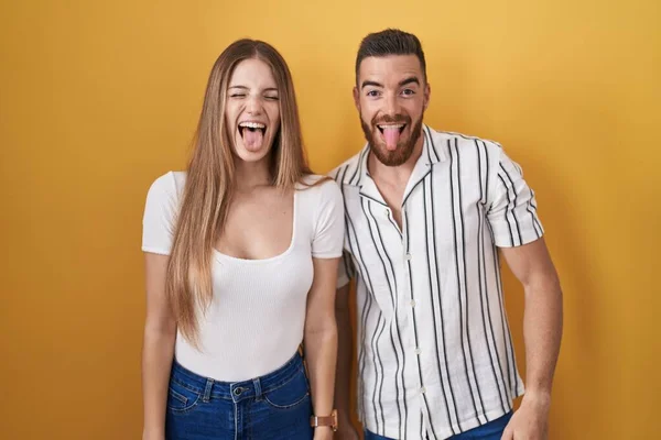 一对年轻夫妇站在黄色的背景上 用滑稽的表情把舌头伸出来 高兴极了 情感概念 — 图库照片