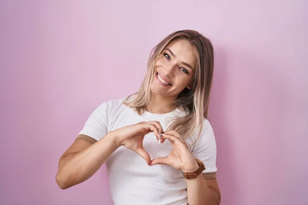 Blonde Caucasian Woman Standing Pink Background Smiling Love Doing Heart — Stockfoto