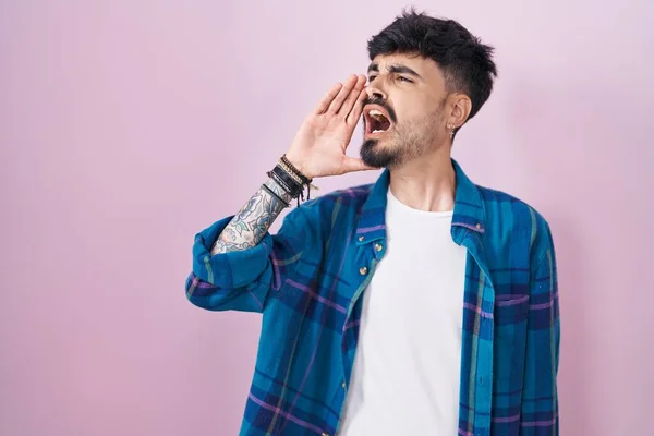 髭を生やした若いヒスパニック系の男がピンクの背景に立って大声で叫び 口の中で手で横に大声で叫ぶ コミュニケーションの概念 — ストック写真