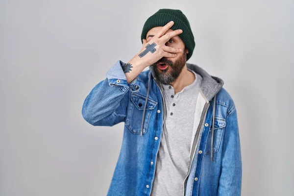 stock image Young hispanic man with tattoos wearing wool cap peeking in shock covering face and eyes with hand, looking through fingers with embarrassed expression. 