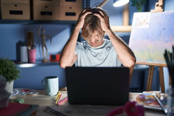 中年男子晚上坐在艺术工作室 带着笔记本电脑 由于疼痛和偏头痛 绝望和压力重重 手放在头上 — 图库照片