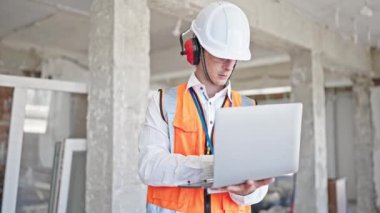 Kafkasyalı genç bir mimar, inşaat alanında dizüstü bilgisayar kullanarak rahat bir ifadeyle duruyor.