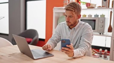 Genç beyaz adam yemek odasındaki masada dizüstü bilgisayar ve akıllı telefon kullanıyor.