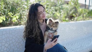 Köpeğiyle gülümseyen genç İspanyol kadın parkta akıllı telefondan selfie çekiyor.