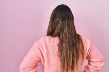 Young hispanic woman standing over pink background standing backwards looking away with arms on body 