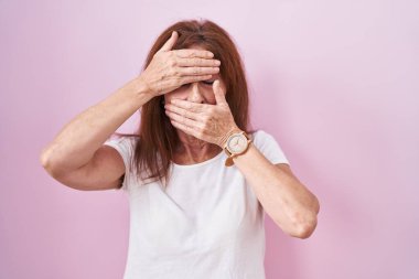 Middle age woman standing over pink background covering eyes and mouth with hands, surprised and shocked. hiding emotion 