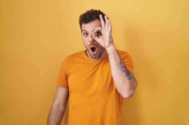 Young hispanic man standing over yellow background doing ok gesture shocked with surprised face, eye looking through fingers. unbelieving expression. 