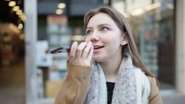 Young blonde woman smiling confident sending voice message by smartphone at street