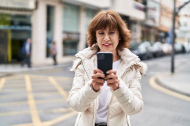Sokakta akıllı telefon kullanan orta yaşlı bir kadın gülümsüyor.