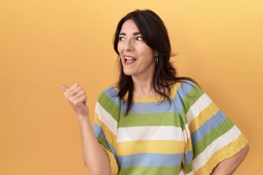 Middle age hispanic woman standing over yellow background smiling with happy face looking and pointing to the side with thumb up. 