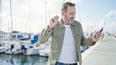 Orta yaşlı bir adam limanda akıllı telefondan müzik dinliyor.