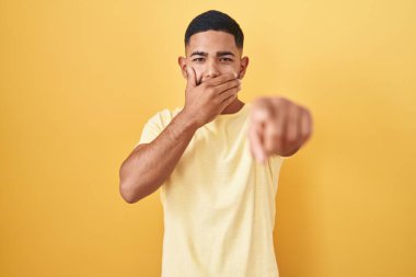 Sarı arka planda dikilen İspanyol genç adam sana gülüyor, eli ağzında kamerayı işaret ediyor, utanç ifadesi. 