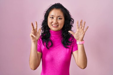 Young asian woman standing over pink background showing and pointing up with fingers number eight while smiling confident and happy. 