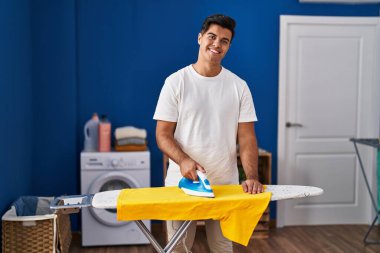 Genç İspanyol adam gülümsüyor çamaşırhanede kendine güvenen ütü yapıyor