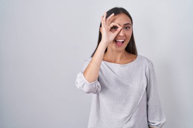 Young hispanic woman standing over white background doing ok gesture with hand smiling, eye looking through fingers with happy face. 