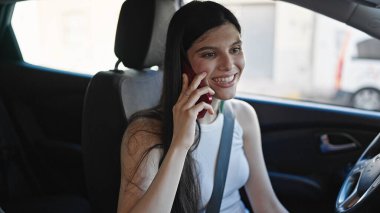 Young beautiful hispanic woman talking on smartphone sitting on car at street