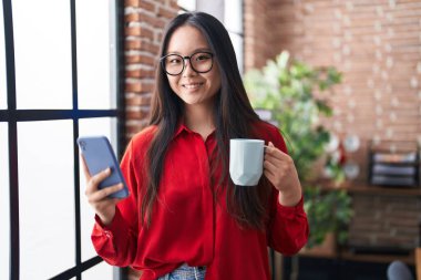 Genç Çinli iş kadını akıllı telefon kullanıyor ofiste kahve içiyor.