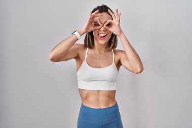 Tecrit edilmiş arka planda spor kıyafetleri giyen İspanyol kadın dürbün gibi jest yapıyor, gözleri parmaklarından bakıyor. çılgın ifade. 