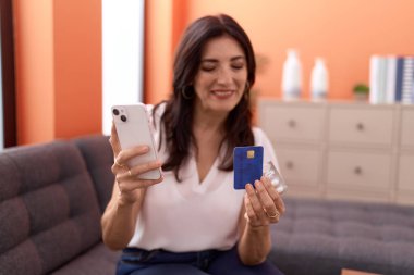 Orta yaşlı İspanyol kadın akıllı telefon ve kredi kartı kullanıyor. Evdeki koltukta oturuyor.