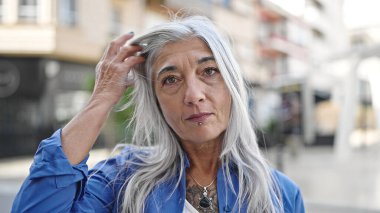 Orta yaşlı gri saçlı kadın ciddi bir ifadeyle ayakta duruyor sokakta saç tarıyor.