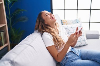 Genç, güzel, İspanyol bir kadın akıllı telefondan video izliyor. Evdeki koltukta oturuyor.